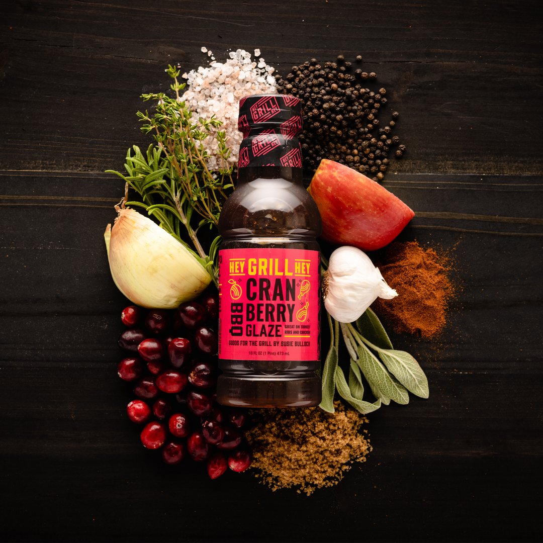 A bottle of Cranberry BBQ Glaze surrounded by select ingredients that include fresh cranberries, onion, garlic, brown sugar, apple, salt, pepper, and other select herbs and spices.