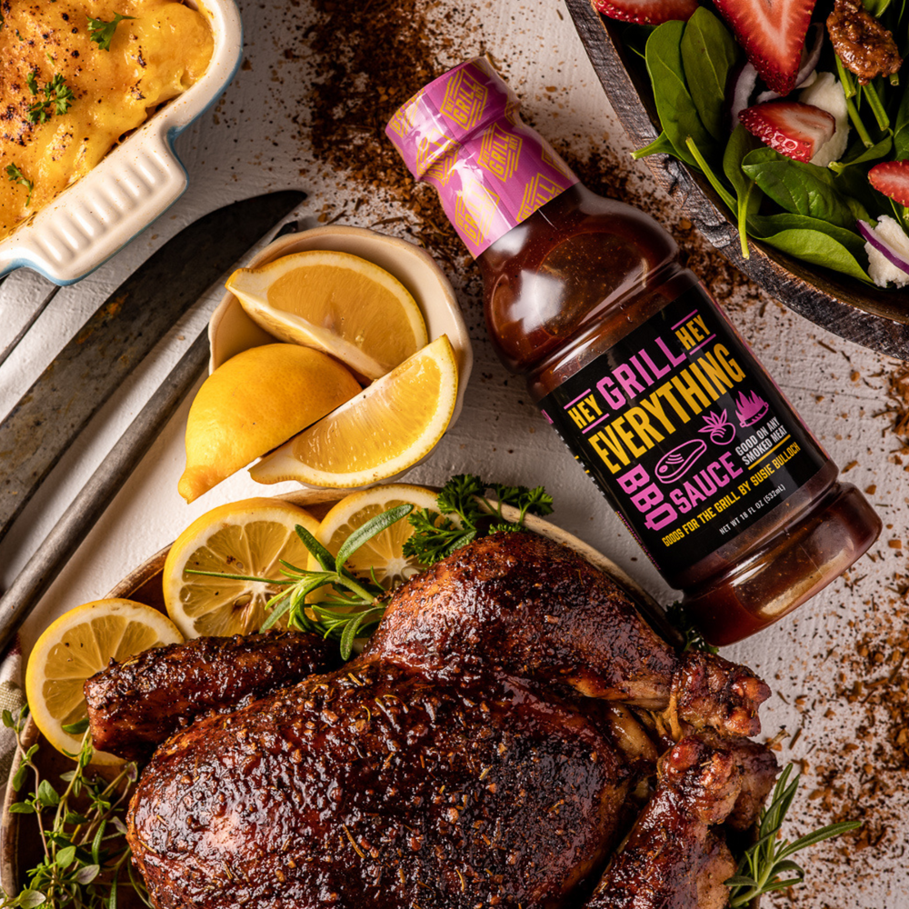 A bottle of Everything Sauce sits on a table next to a smoked whole chicken that has been plated with herbs and lemon. Other side dishes surround the area