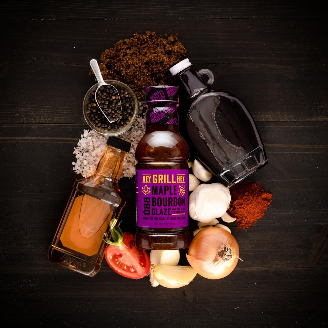 A bottle of Maple Bourbon BBQ Glaze surrounded by ingredients including, maple syrup, a bottle of bourbon, a tomato, an onion, garlic, brown sugar, and other select spices.