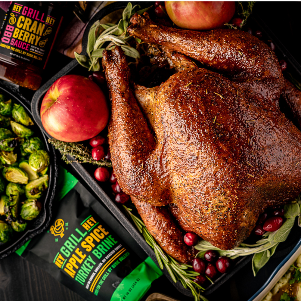 A beautiful smoked turkey sits plated with fresh herbs, cranberries, and apples. Next to it is a bottle of Cranberry glaze and Apple Spice Turkey Brine.