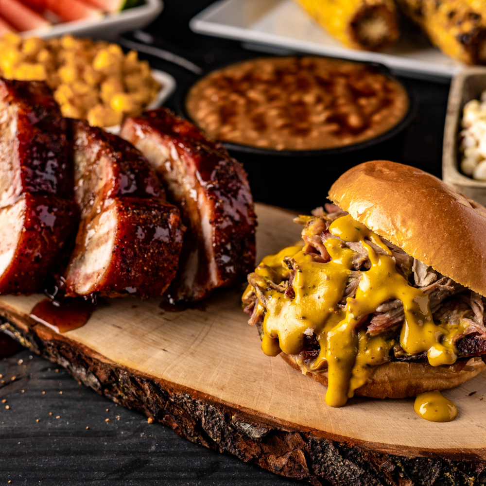 Ribs covered in maple bourbon glaze and a pulled pork sandwich with zesty gold sauce, plated with other side dishes in the background. 