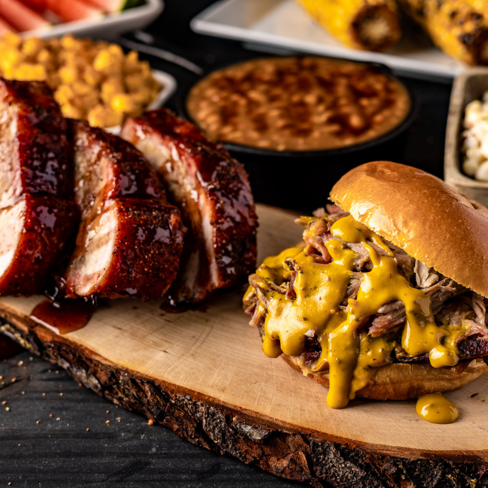 Photo of pulled pork on a toasted bread bun, and the pork is smothered in Hey Grill Hey Zesty Gold Sauce. The pulled pork sandwich is on a wooden board next to three sauced up pork ribs.