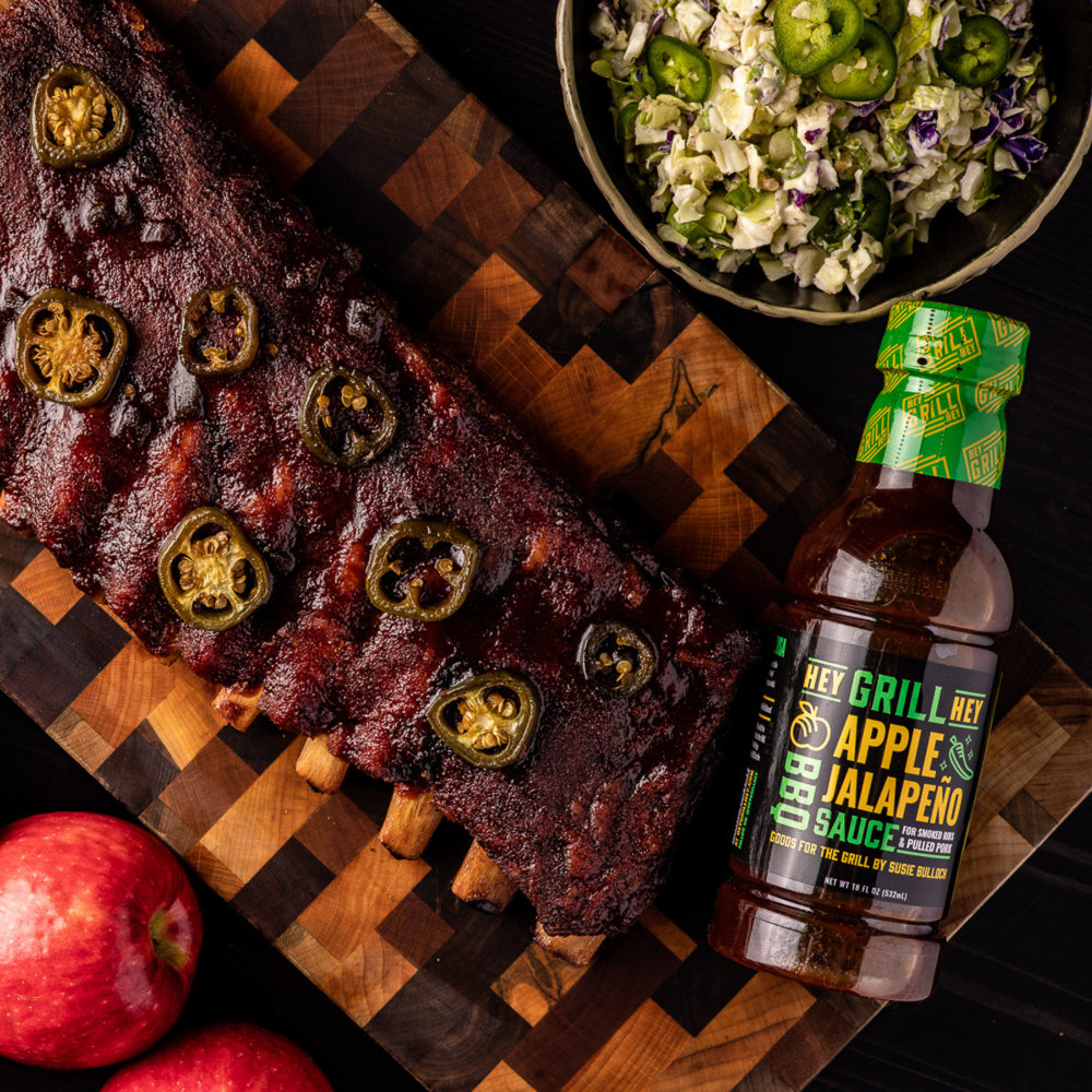 A bottle of apple jalapeno bbq sauce on a cutting board with a rack of jalapeno smoked ribs. Coleslaw and apples sit on the side of the cutting board. 