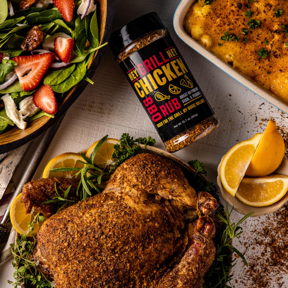 A bottle of Hey Grill Hey Chicken rub lying on a table and surrounded by three food dishes: A whole smoked chicken that was seasoned with Hey Grill Hey Chicken Rub, a dish of Smoked Macaroni and Cheese, and a bowl of green salad garnished with sliced strawberries.