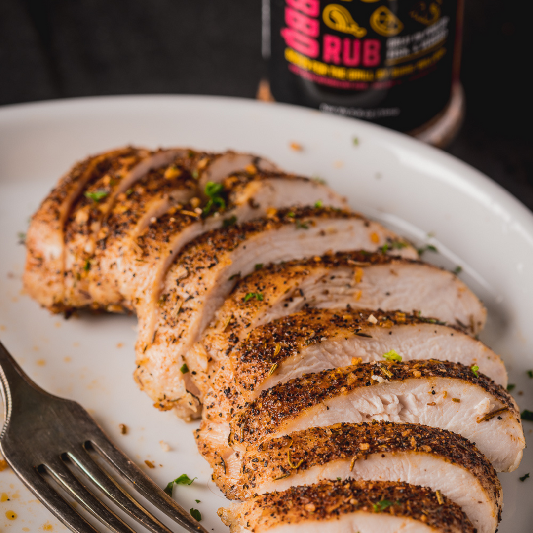 A close up shot of a  sliced, grilled chicken breast on a white plate with a bottle of Hey Grill Hey Chicken Rub behind it.