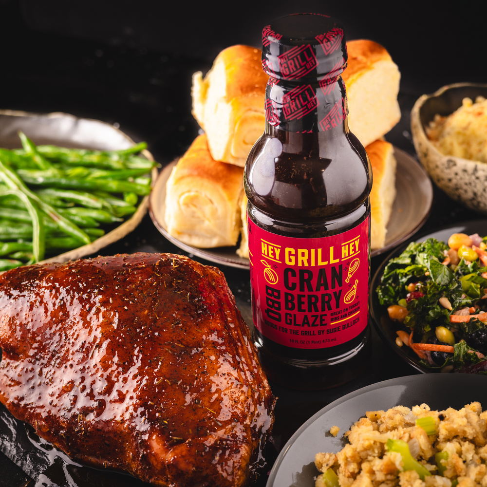 A bottle of Cranberry BBQ Glaze next to a glazed, roasted turkey breast. Other side dishes including a salad, rolls, and beans are on the table. 