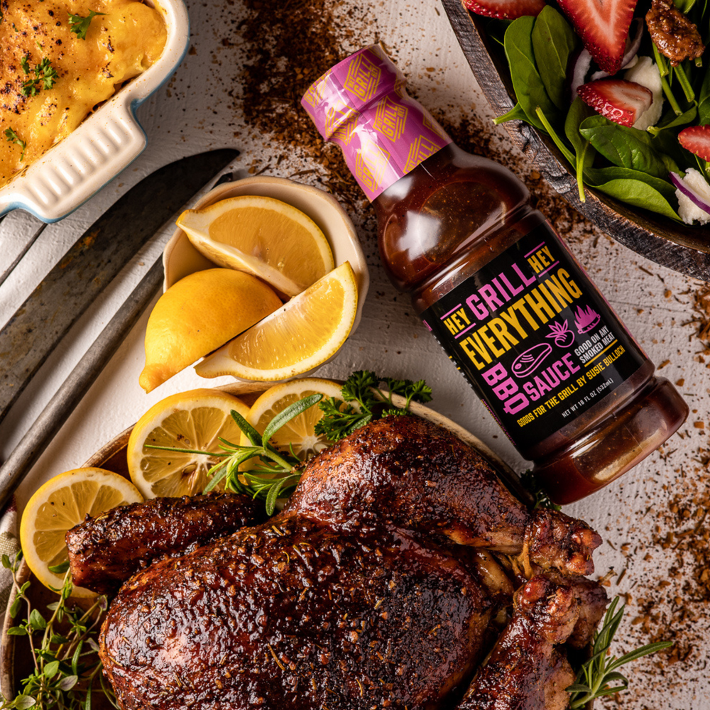 A bottle of Everything Sauce sits on a table next to a smoked whole chicken that has been plated with herbs and lemon. Other side dishes surround the area 