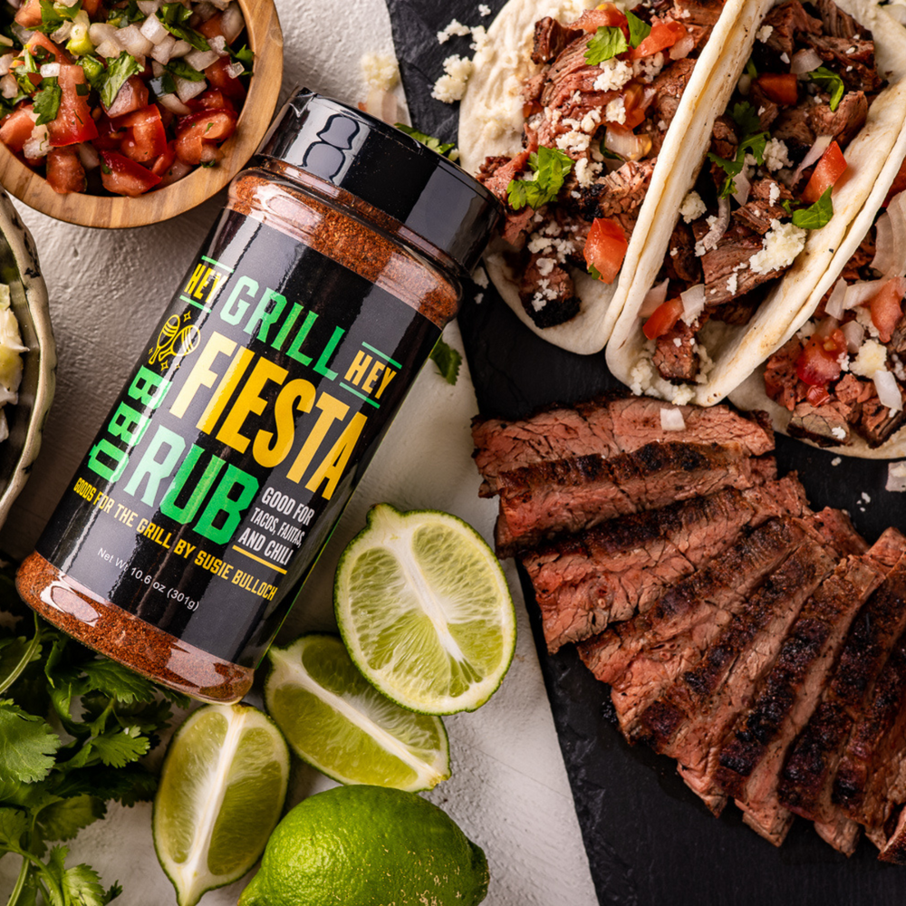 A bottle of Hey Grill Hey Fiesta rub laying on a table, and surrounded by three steak tacos, sliced steak, sliced limes, cilantro, and a small bowl of fresh salsa.