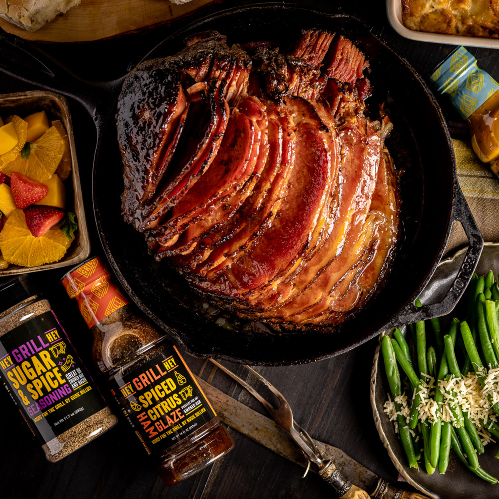 Overhead shot of a smoked spiral-cut ham on a cast iron skillet. Next to the ham are the Hey Grill Hey Sugar and Spice Seasoning, the Hey Grill Hey Spiced CItrus Ham Glaze, and Hey Grill Hey Zesty Gold BBQ Sauce.