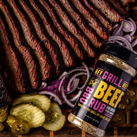 Sliced Smoked Brisket resting on a wooden cutting board next to sliced pickles, sliced red onions, and a bottle of Hey Grill Hey Beef Rub.