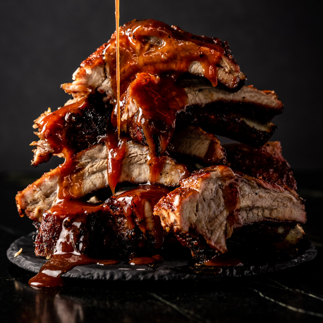 Sliced ribs piled up high on a black plate, with BBQ sauce drizzling down on top of the pile.