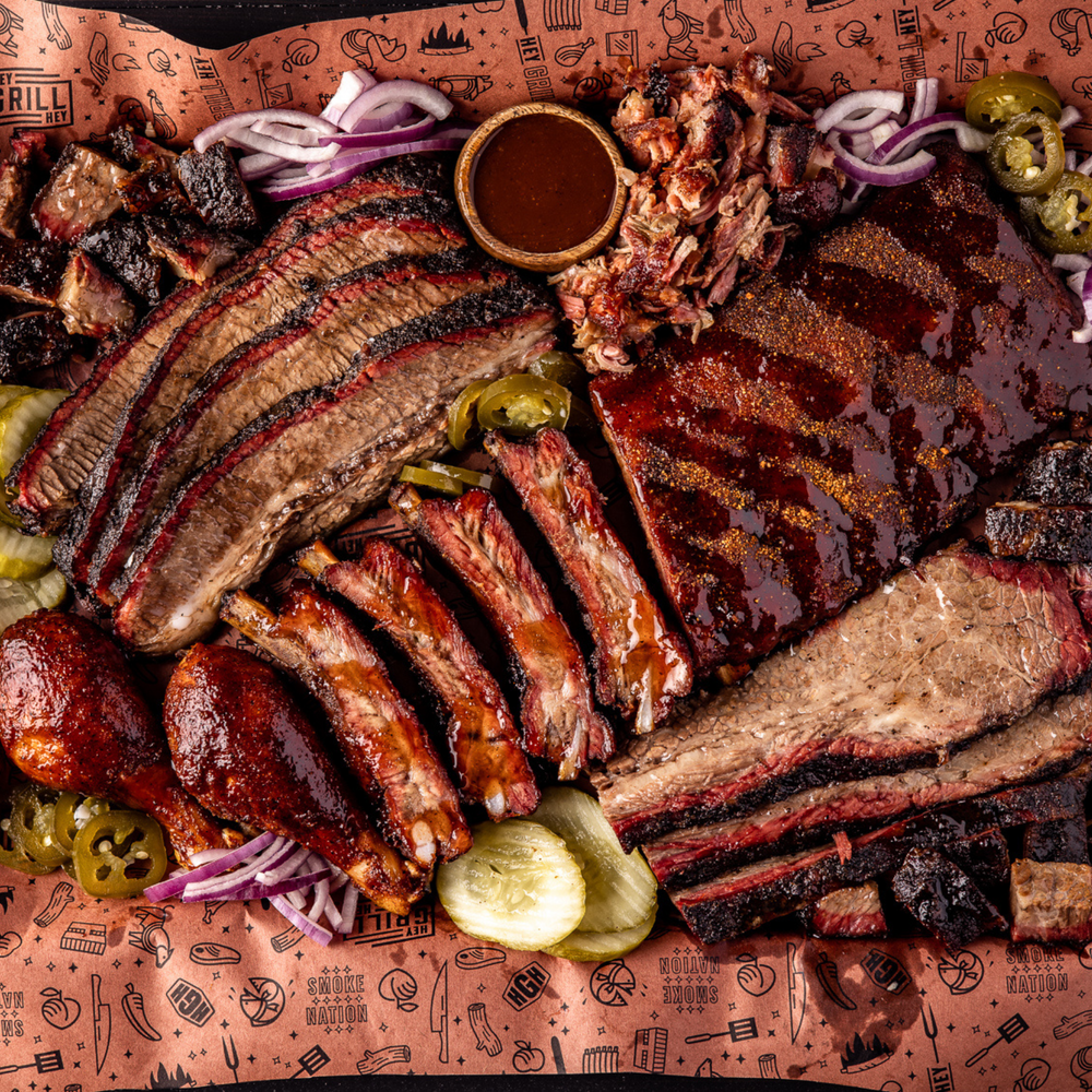A large platter with butcher paper covered with cooked and sliced brisket, cooked chicken drumsticks, cooked pork ribs, and garnished with sliced pickles, sliced jalapenos, and sliced red onions,