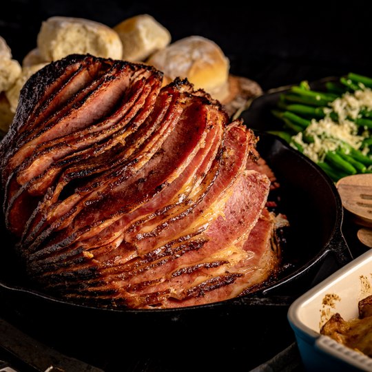 A smoked, glazed ham that is spiral cut, sitting in a cast iron pan with other side dishes in the background. 