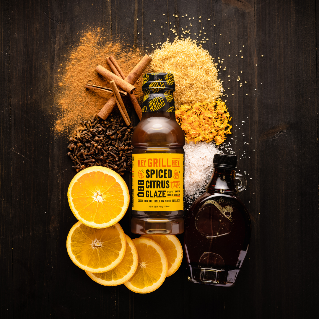 A bottle of Spiced Citrus BBQ Glaze surrounded by ingredients including sliced oranges, cinnamon, maple syrup, salt, brown sugar, and other select spices.   