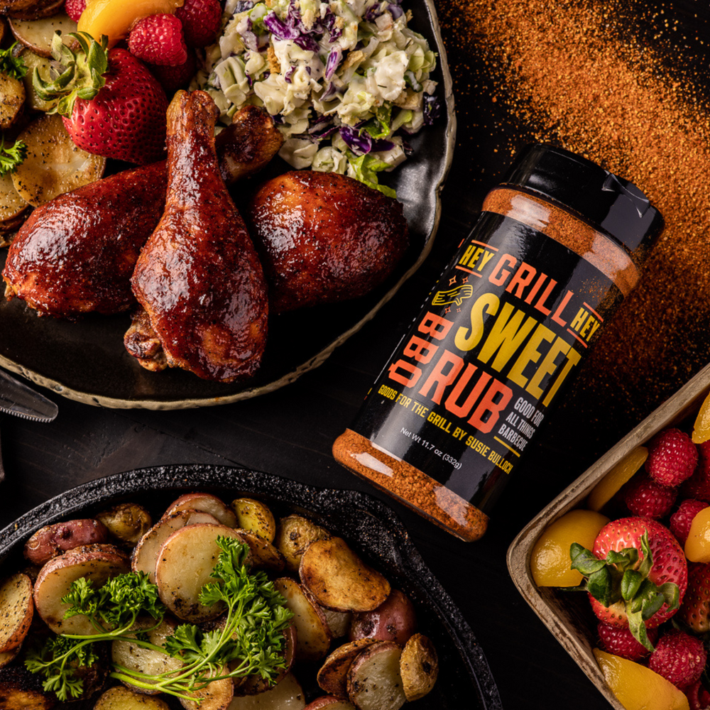 A bottle of Hey Grill Hey Sweet Rub lying on a table surrounded by a plate of grilled chicken drumsticks, a cast iron skillet with sliced potatoes, and a glow of strawberries.