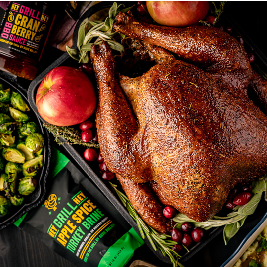 A beautiful smoked turkey sits plated with fresh herbs, cranberries, and apples. Next to it is a bottle of Cranberry glaze and Apple Spice Turkey Brine.  