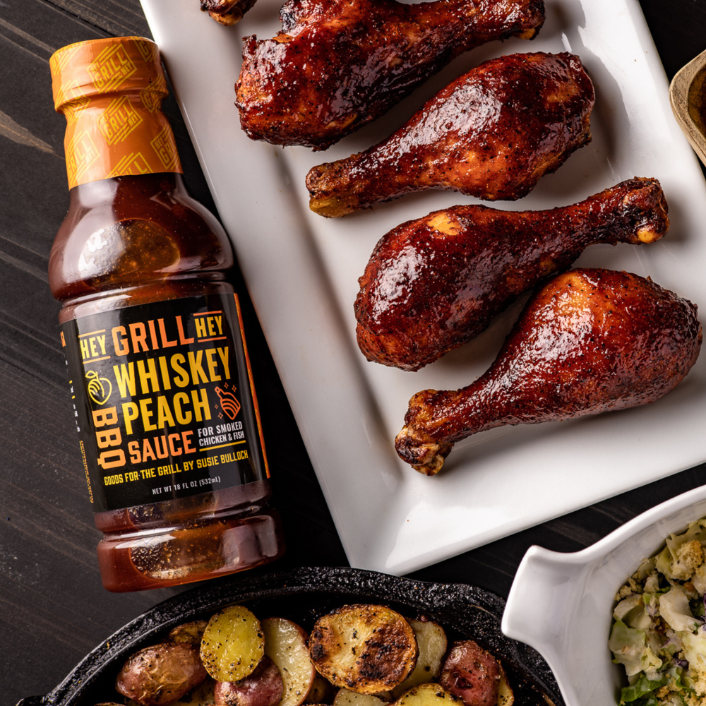A bottle of Whiskey Peach BBQ Sauce sits on a table next to smoked chicken legs covered in BBQ Sauce. 