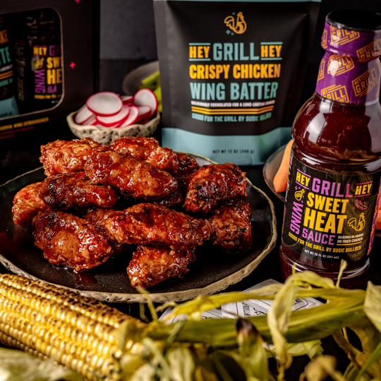 A plate of smoked and friend chicken wings on a plate, next to a bottle of Hey Grill Hey Sweet Heat Wing Sauce and a bag of the Hey Grill Hey Crispy Chicken Wing Batter.
