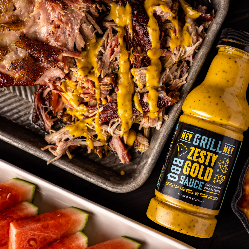 A bottle of Zesty Gold BBQ Sauce sits next to a pile of smoked pulled pork and drizzled with the yellow Zesty Gold BBQ Sauce. 