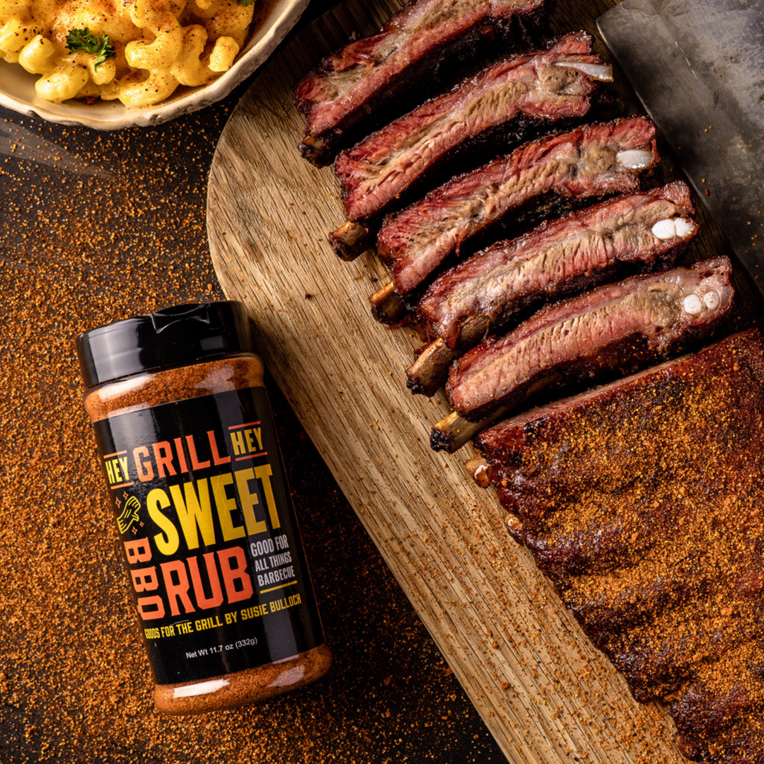 An overhead shot of smoked and sliced ribs on a wooden cutting board, with a bottle of Hey Grill Hey Sweet Rub next to it.